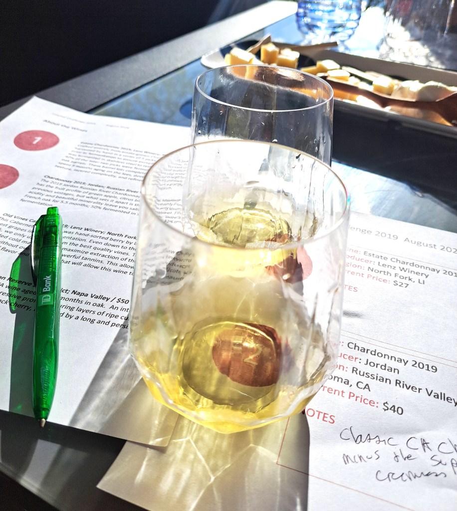 Two glasses of white wine on a table, accompanied by a green pen and a sheet of paper with tasting notes and pricing information.