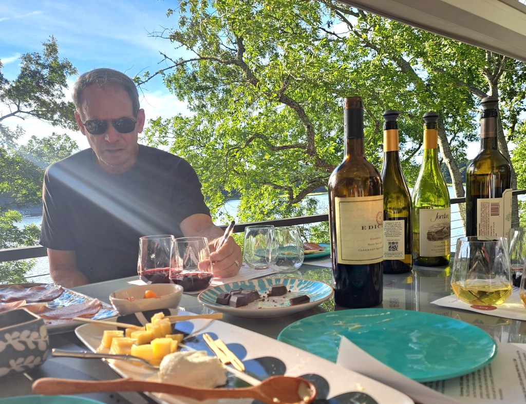 A man sitting at a table with various food items and several wine bottles, enjoying a scenic view of greenery and water.